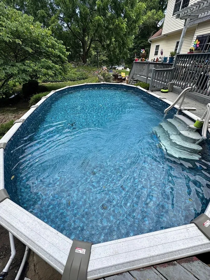 Backyard pool with blue water, white deck, and house patio beside lush green trees