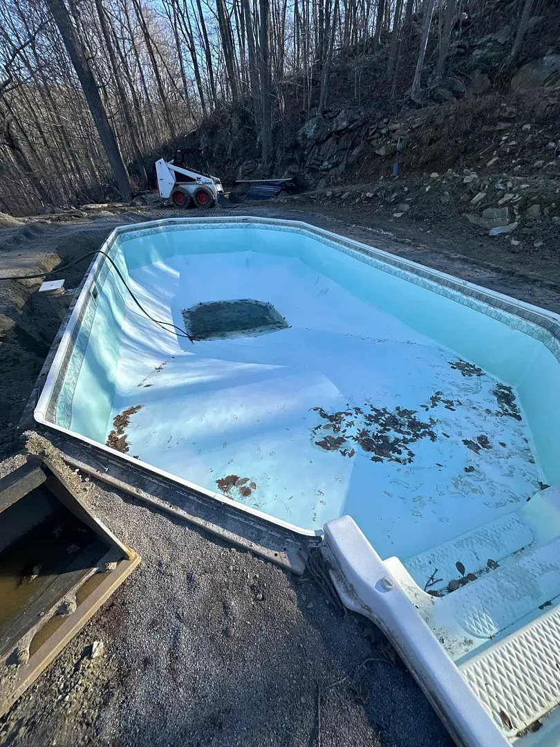 Empty outdoor pool with cracked liner and debris, surrounded by leafless trees and a rocky slope