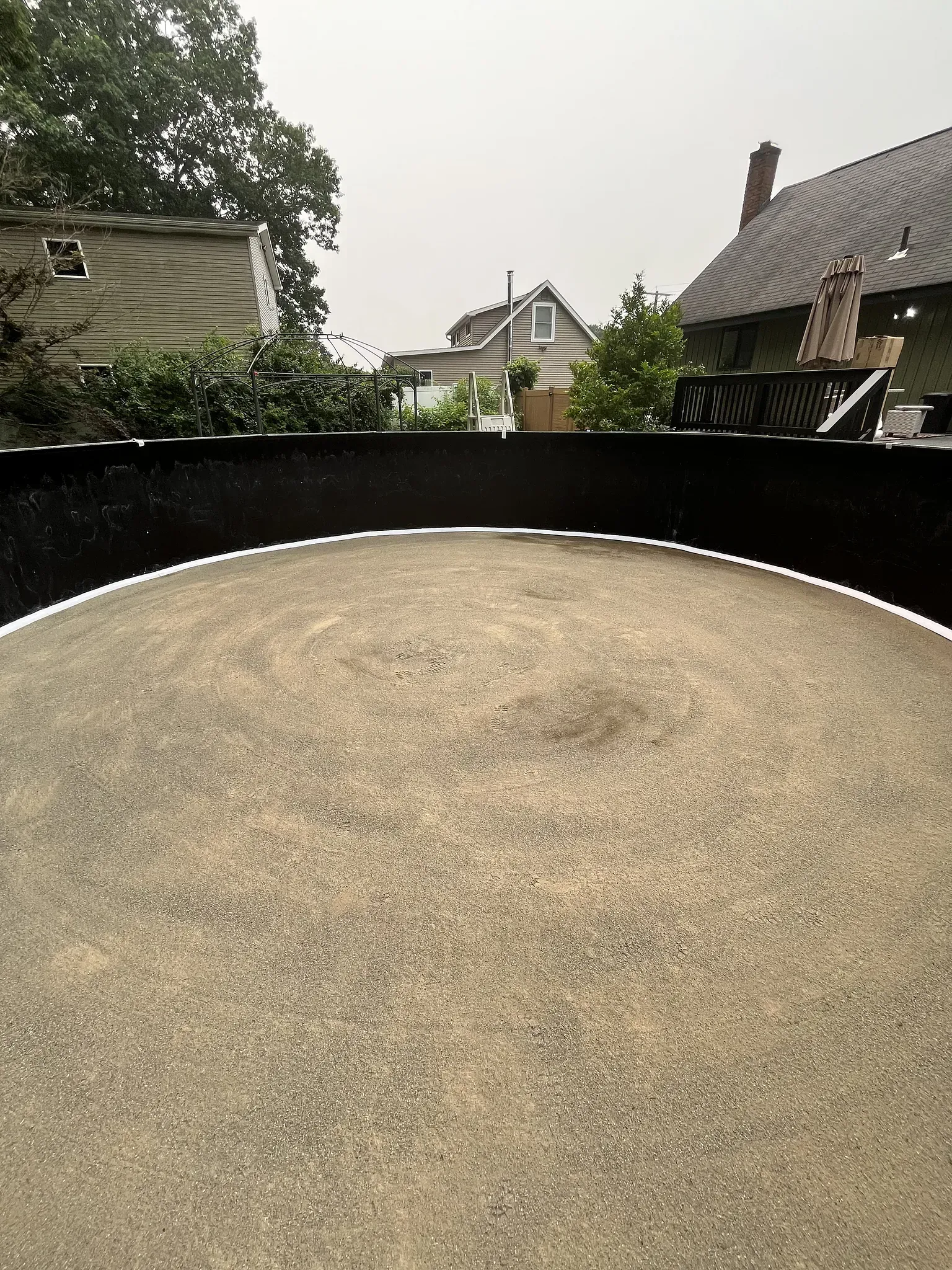 Circular gravel surface in a backyard, with black edging and houses visible beyond greenery under a cloudy sky