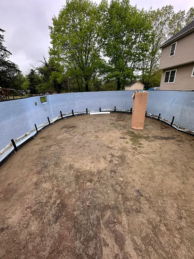 Curved blue retaining wall around a bare dirt yard with a brick column and trees in the background