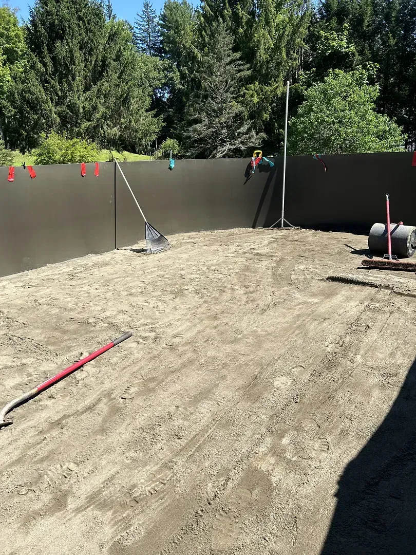 Backyard construction site with leveled dirt, dark fence, tools, and trees in the background