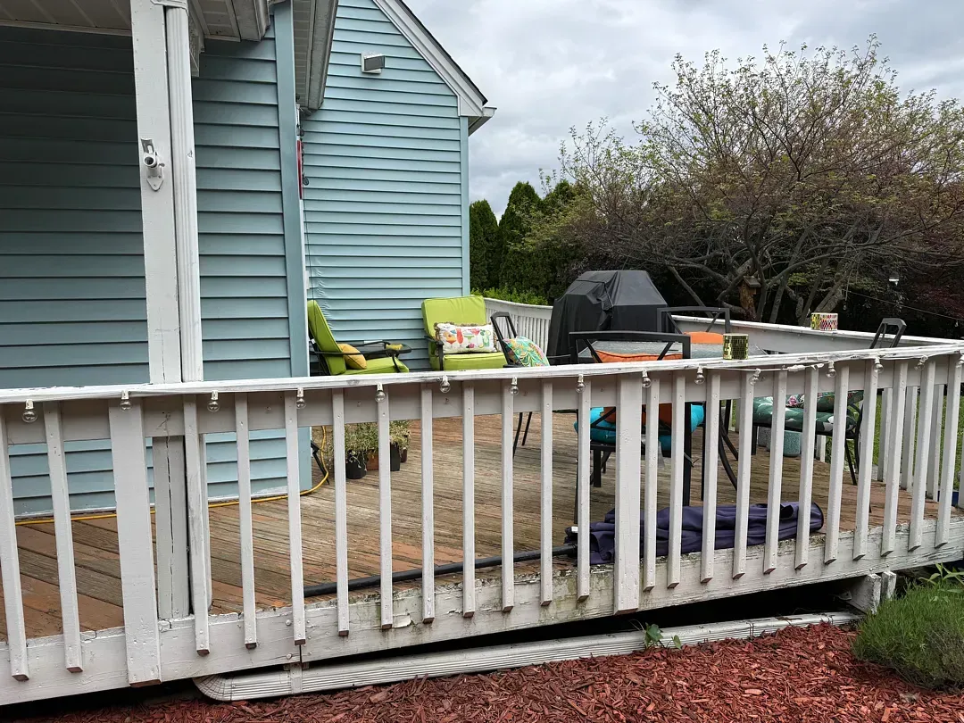 White porch railing in front of a light blue house with patio chairs and a grill on a gravel yard.