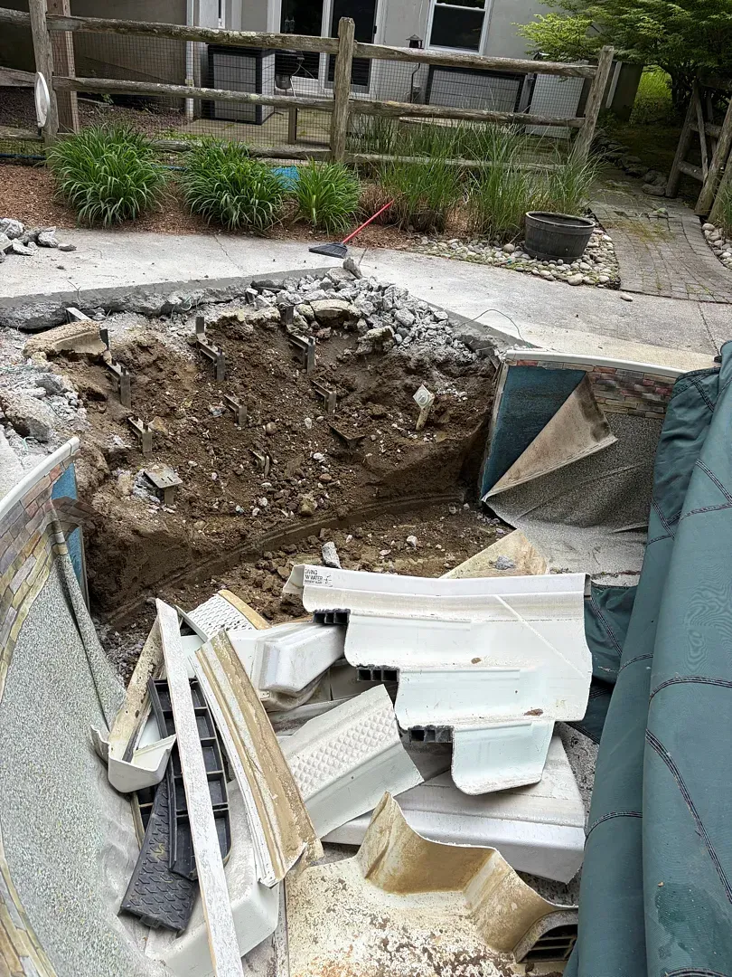 Backyard excavation hole beside a patio, with broken white debris and garden plants nearby