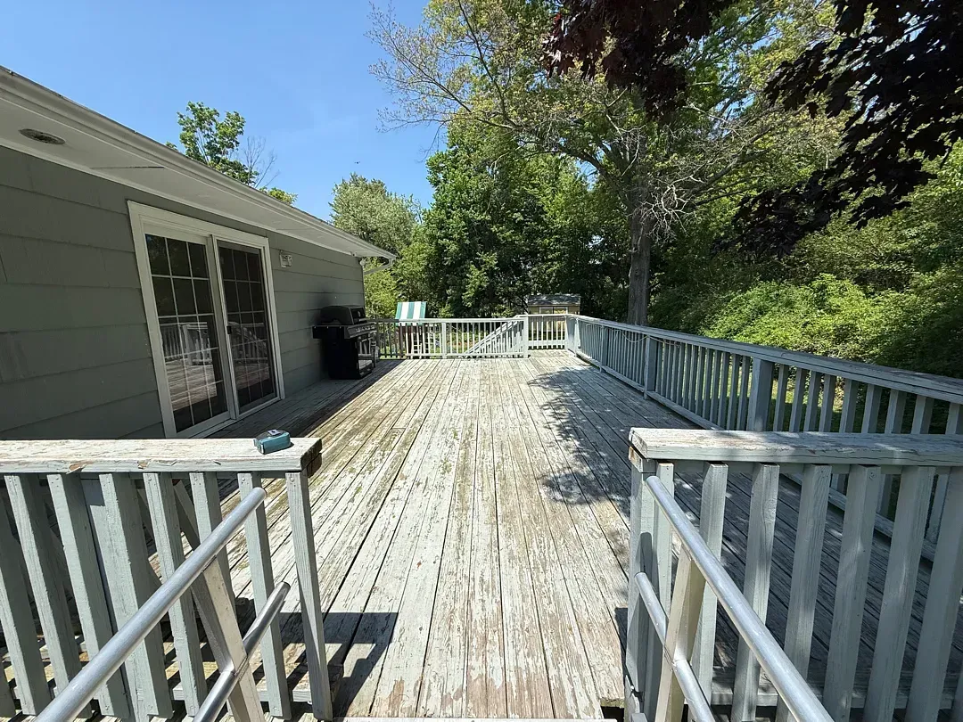 Wooden deck with white railings beside a house, leading toward trees in bright sunlight