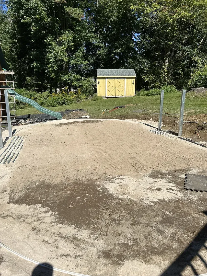 Freshly graded dirt area with posts and a yellow shed in a wooded backyard