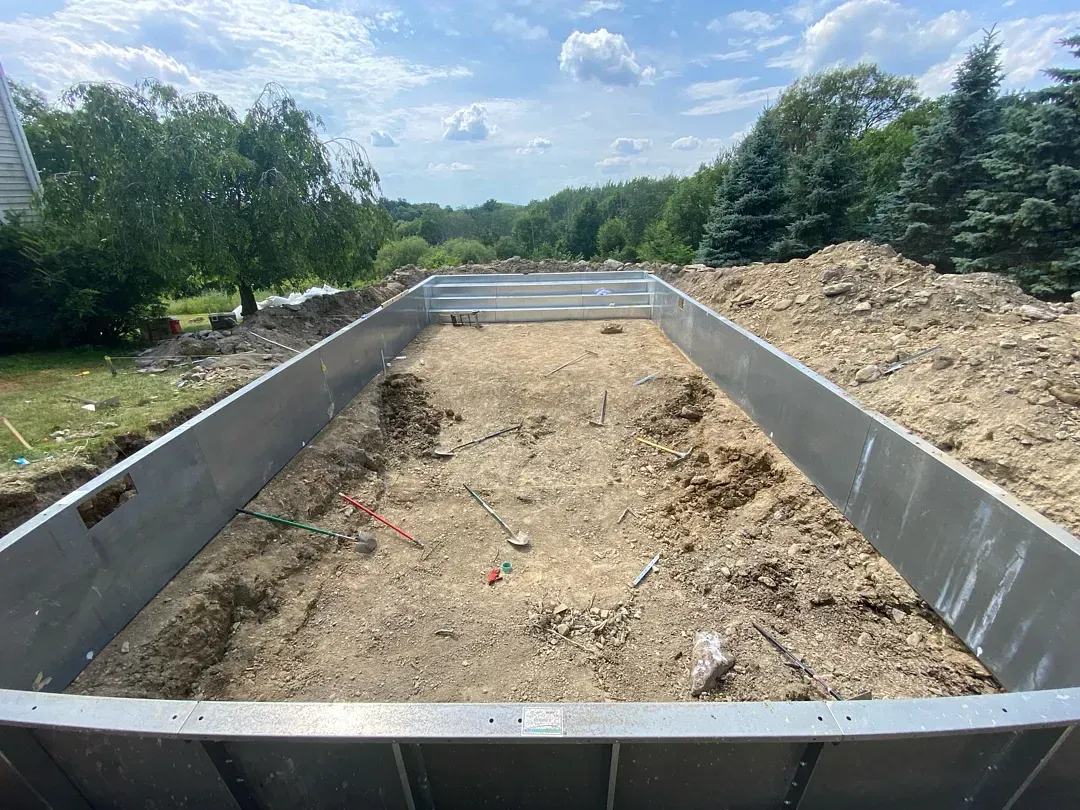 Concrete foundation trench under construction, with gravel and dirt between gray retaining walls on a sunny day