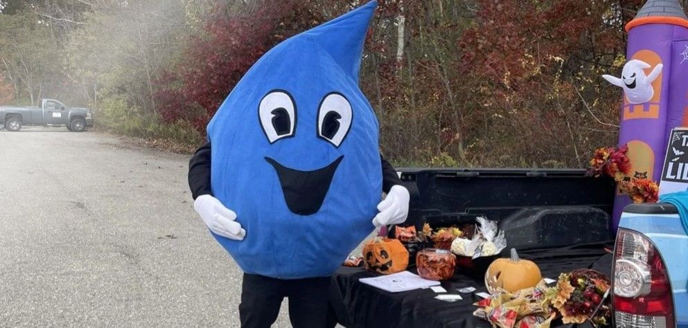 Blue water-drop mascot standing by a pickup truck tailgate with food and drinks at an outdoor event