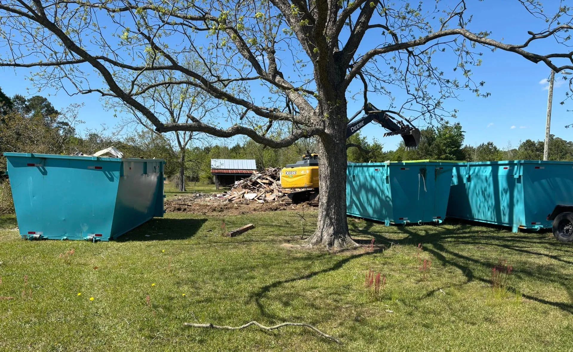 Three teal dumpsters with an excavator near a tree on a grassy field; demolition in progress.