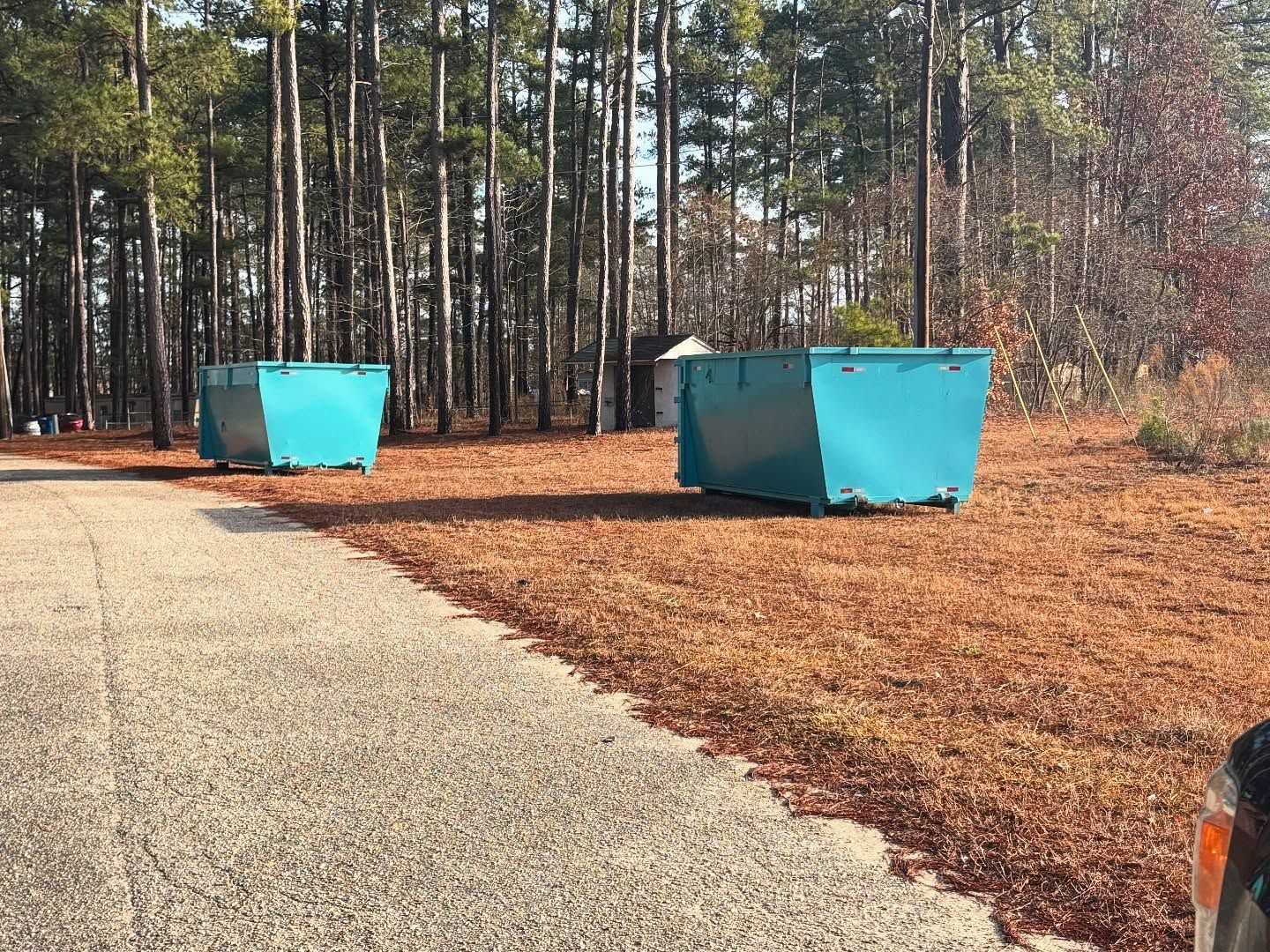 Two large teal dumpsters on a brown-needle-covered ground, near a paved road and a forest.