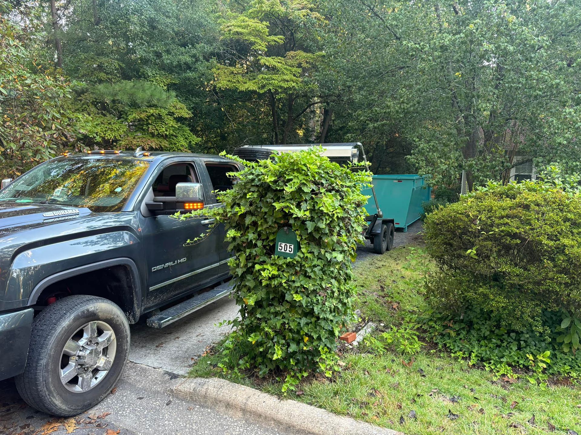 Dark gray pickup truck with a green trailer on a driveway, surrounded by green foliage.