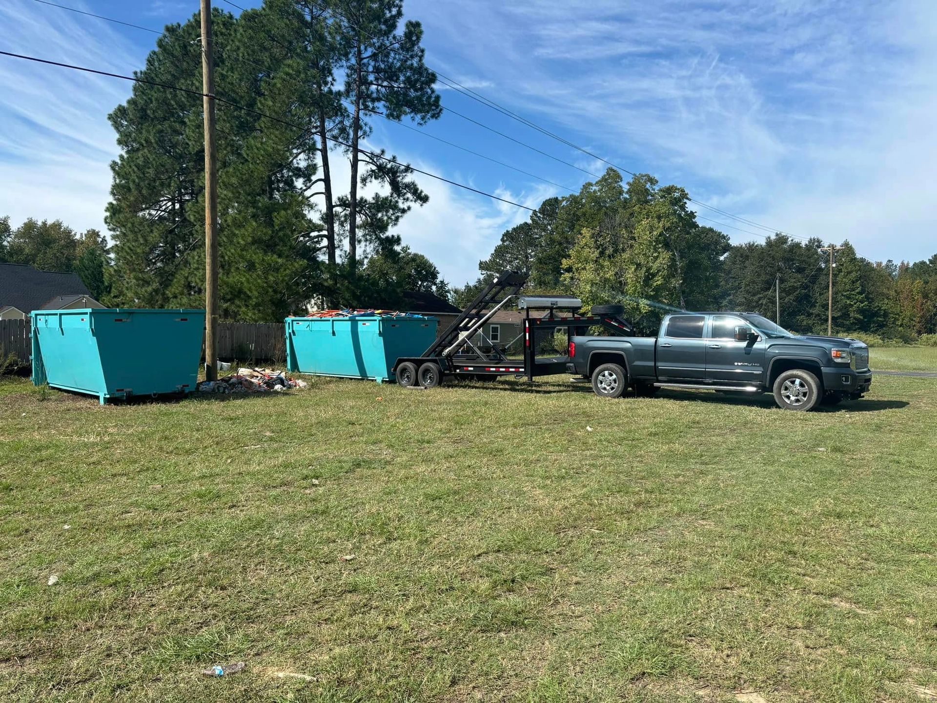 Two teal dumpsters and a trailer hitched to a gray pickup truck on a grassy field under a blue sky.