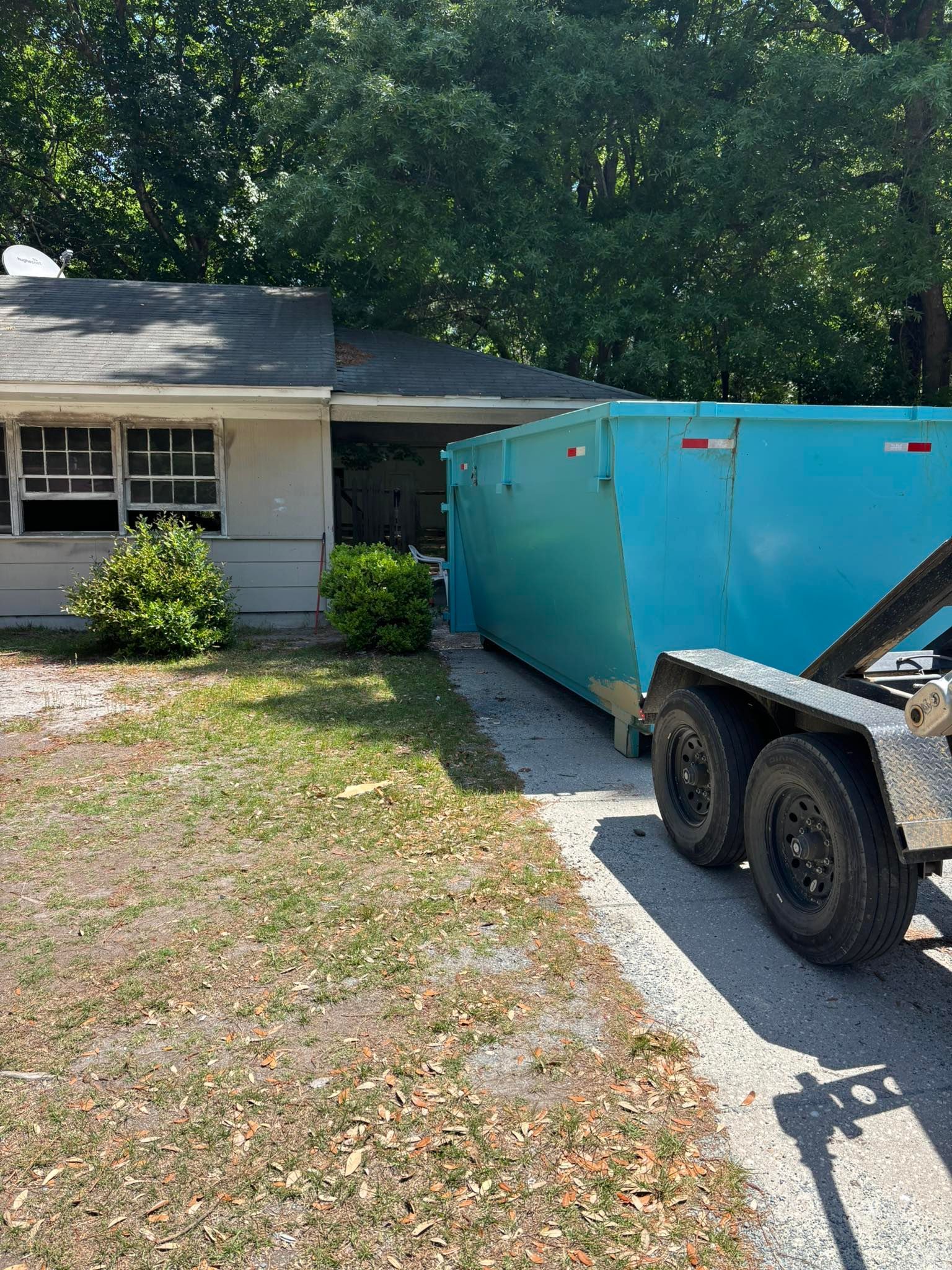 Blue dumpster on a trailer parked next to a light-colored house with a small front yard.
