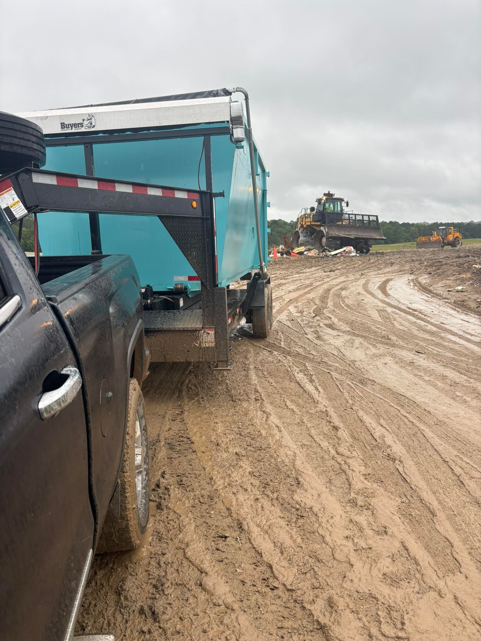 Black truck towing a teal dump trailer on a muddy construction site, heavy machinery in the background.