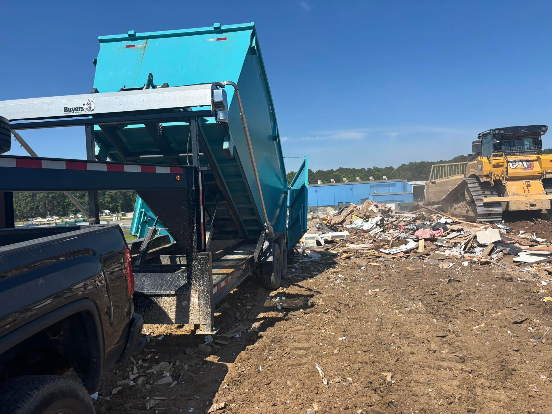 A teal dumpster being emptied at a landfill. A bulldozer pushes trash.
