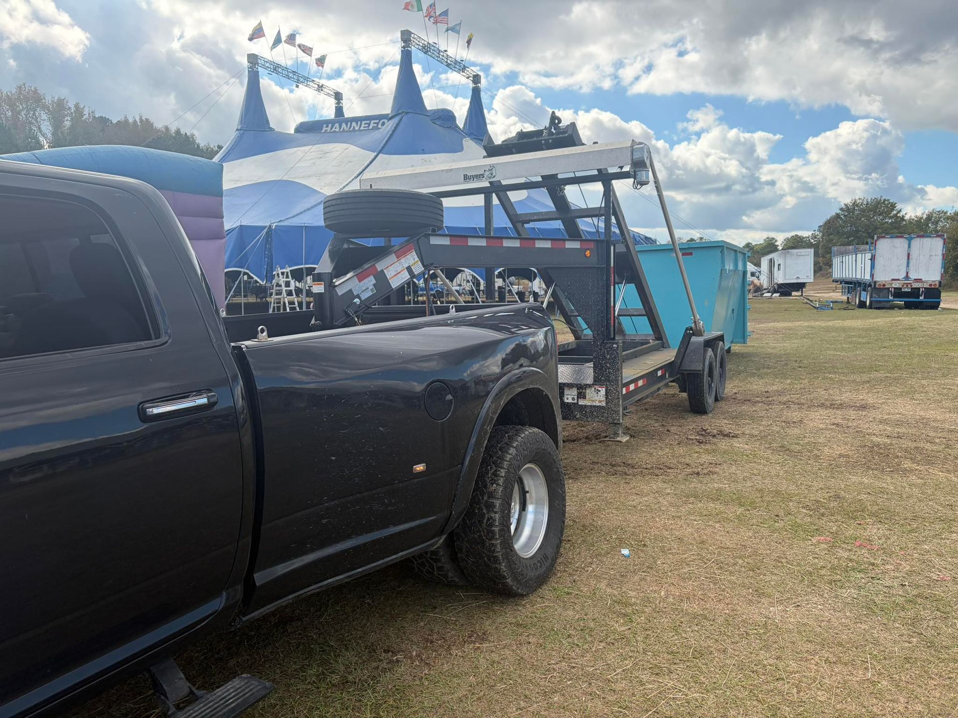Black truck towing a blue dumpster trailer on a grassy field; circus tent in the background.