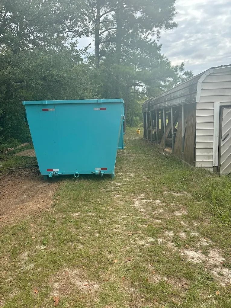 A teal dumpster sits on grassy ground next to a weathered wooden building and trees under a cloudy sky.