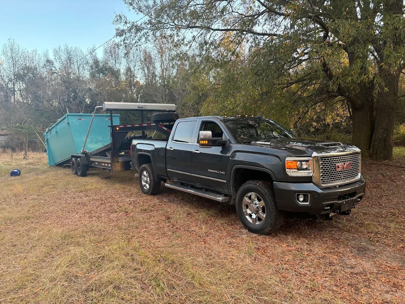 Dark gray pickup truck towing a teal dumpster trailer on a grassy field with trees.