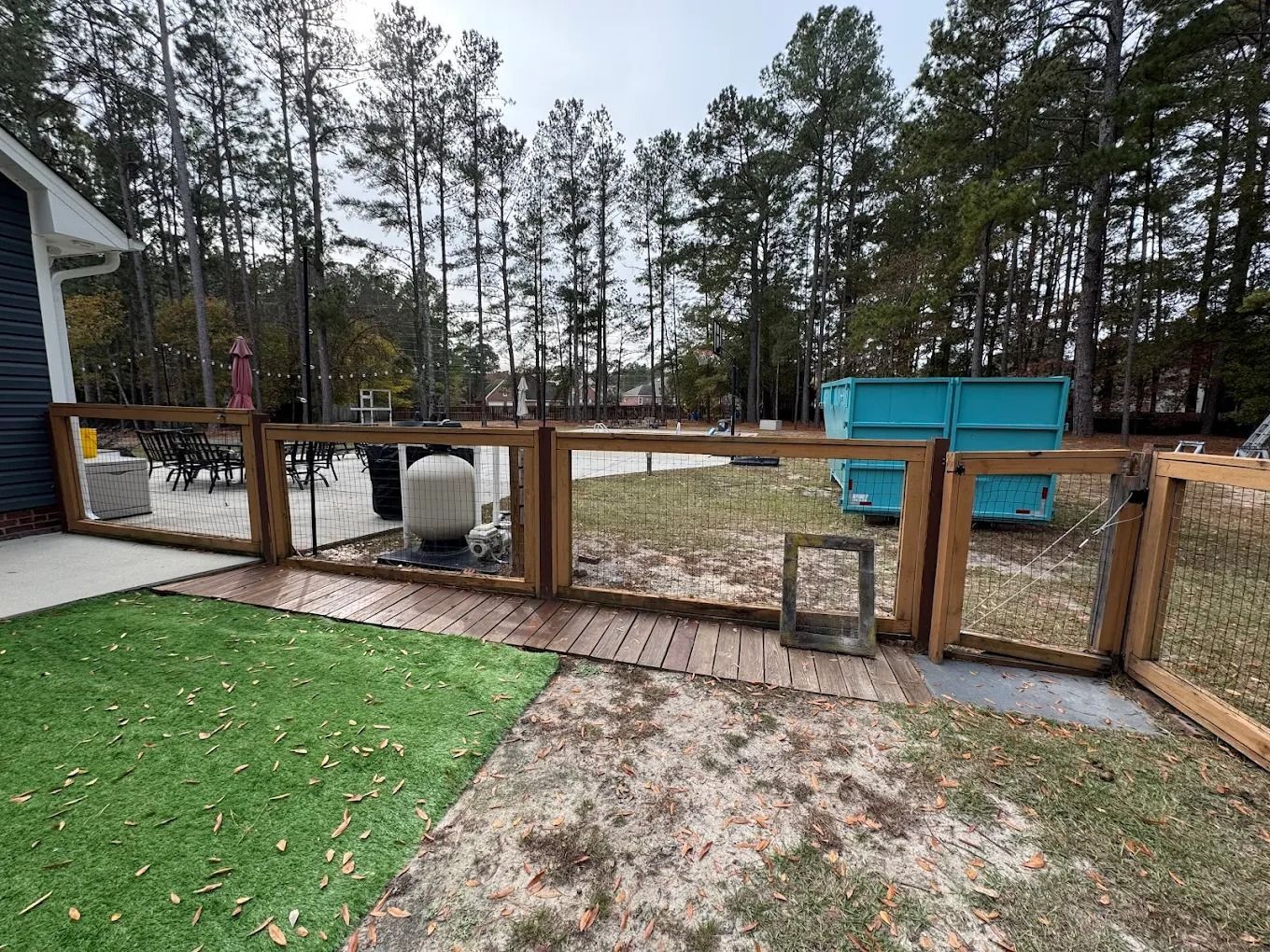 Wooden fence with wire mesh encloses a backyard with green grass, sand, and blue dumpster.