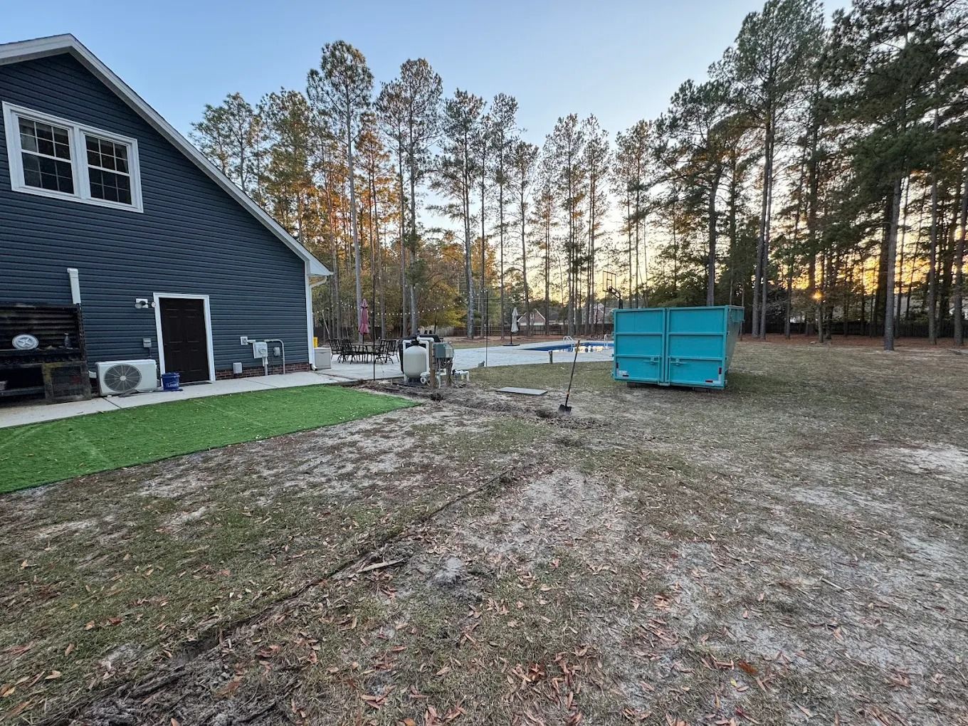 Blue house with yard, teal dumpster. Trees in background. Grass and dirt.