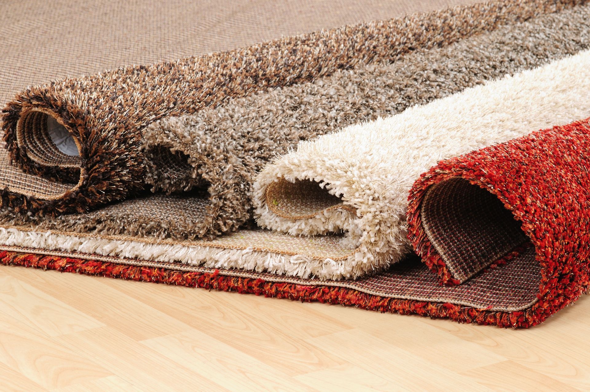 Rolled area rugs in brown, beige, and red stacked on a wooden floor.