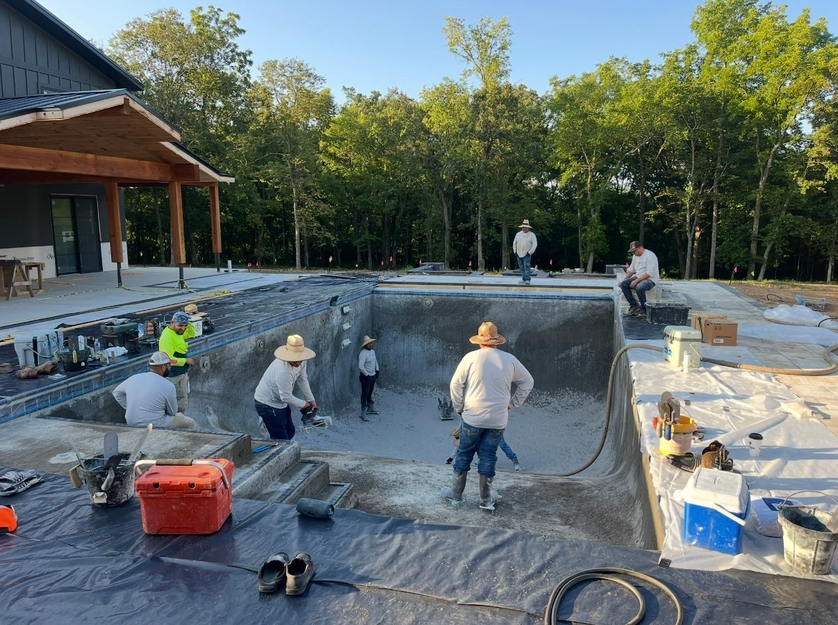 Workers constructing a pool. Several people are in the pool working. House and trees in the background.