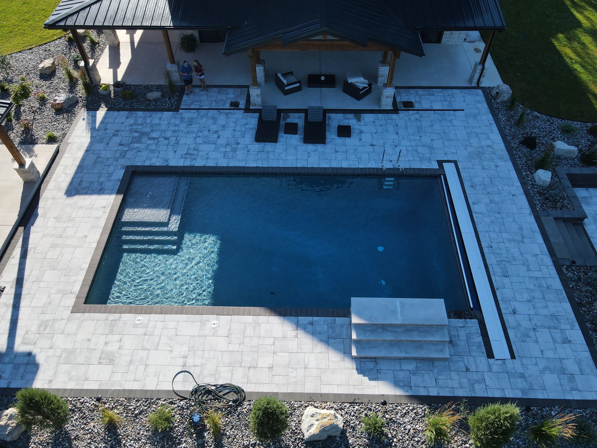 Aerial view of a rectangular pool with stone patio, surrounded by landscaping and a covered outdoor seating area.