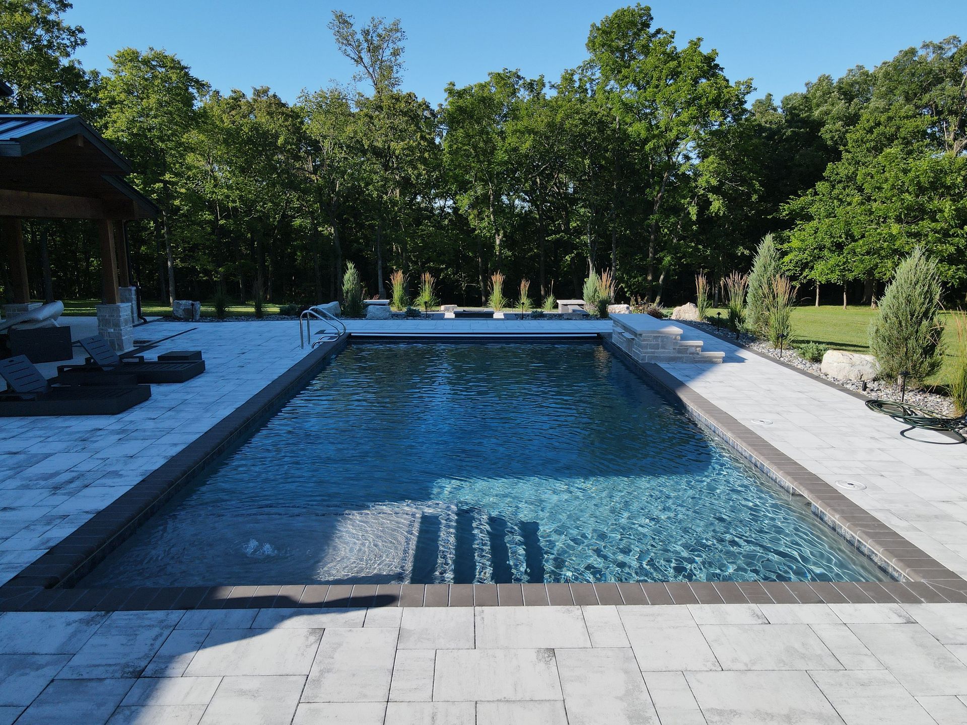 Rectangular pool with steps, paved patio, and trees in background.