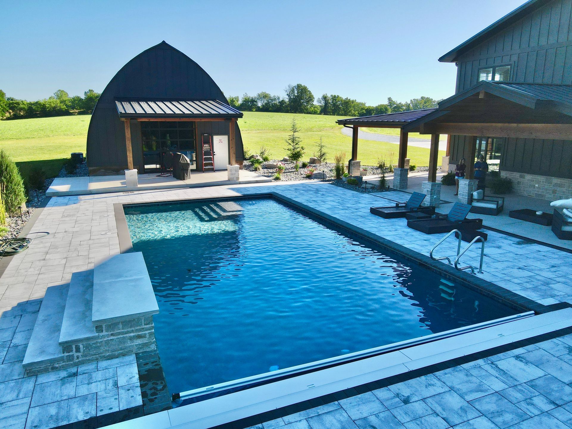 Pool with stone steps, a house, and a unique arched structure in a sunny outdoor setting.