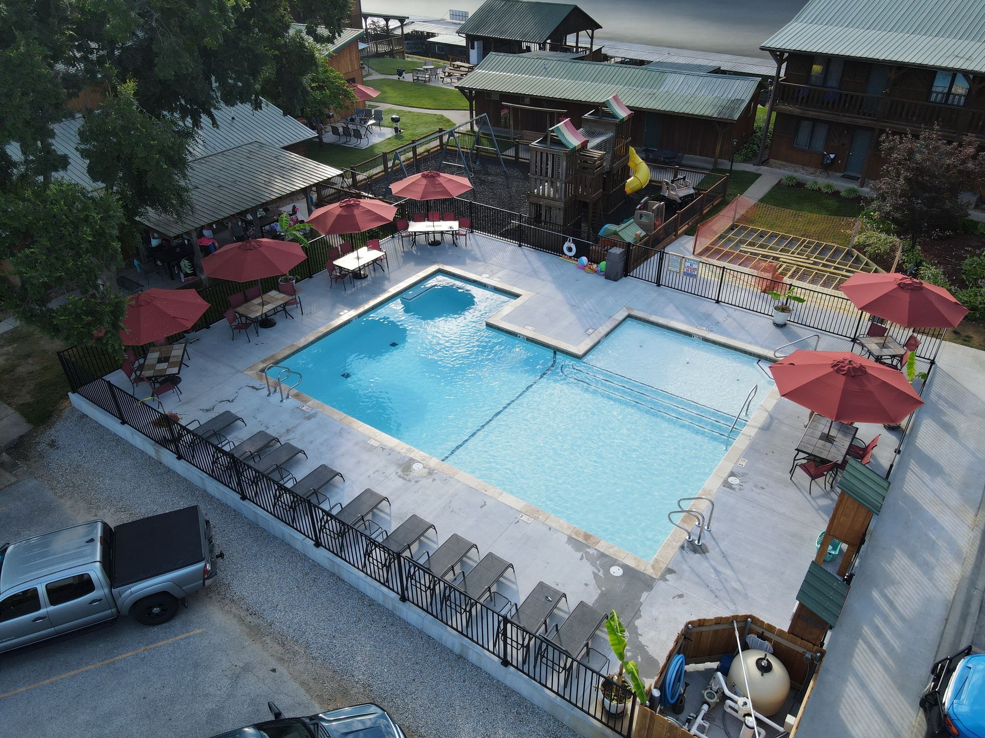 Overhead view of a swimming pool surrounded by red umbrellas, tables, lounge chairs, and buildings.