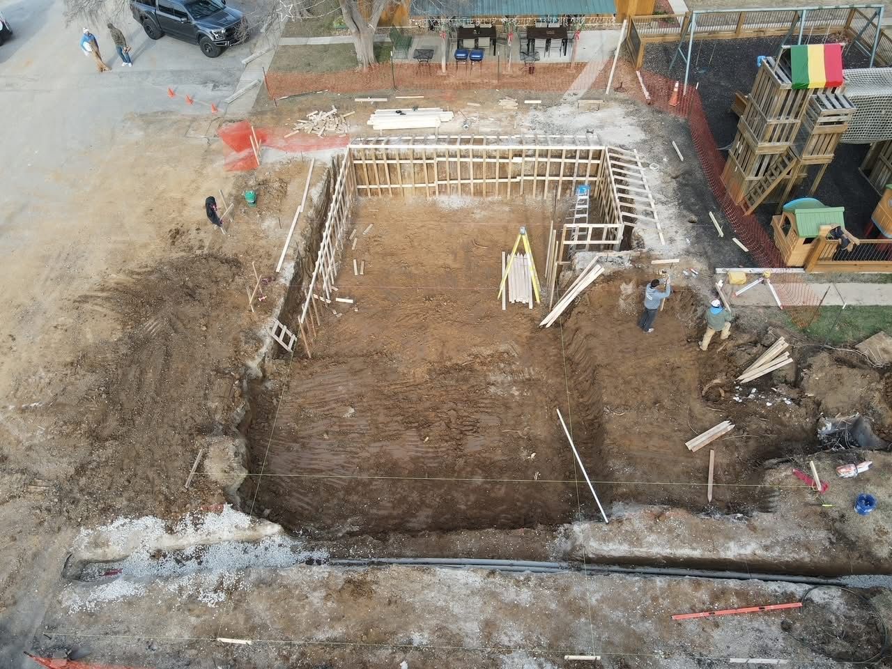 Construction site: excavation for a building foundation, with wooden framing, workers, and a car.