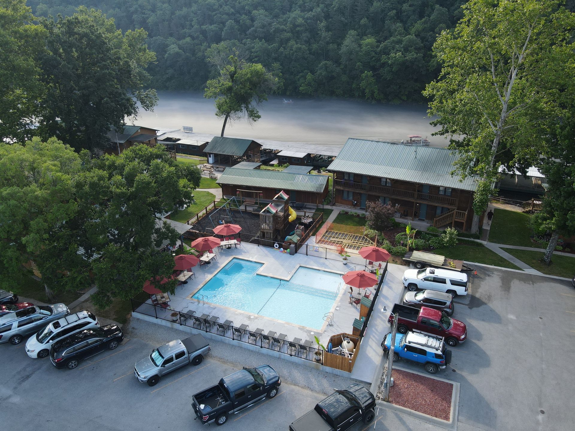 A resort with a pool, red umbrellas, and parked cars near a river. Lush greenery and buildings are in the background.