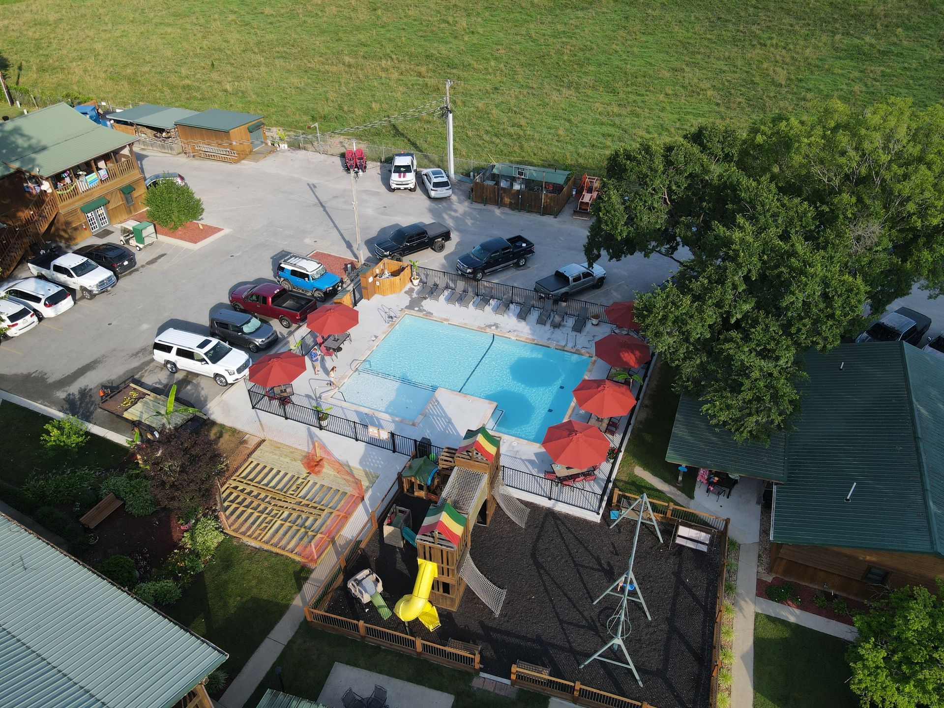 Aerial view of a resort with a pool, playground, parking lot, and cabins in a green, grassy area.