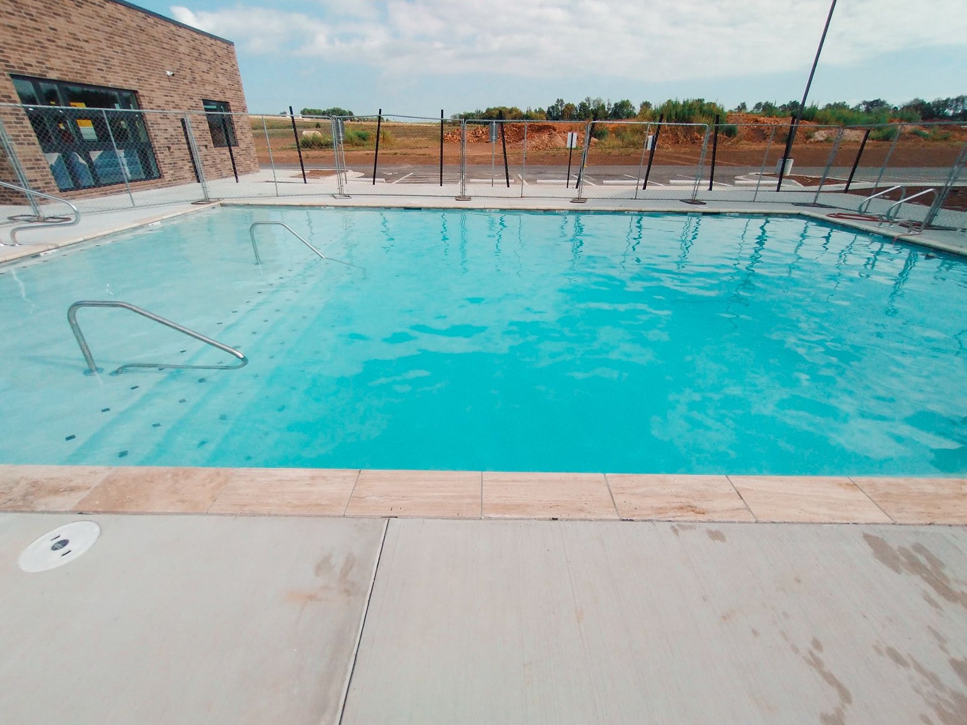 Outdoor swimming pool on a sunny day. Light blue water, gray concrete deck, and a brick building in the background.