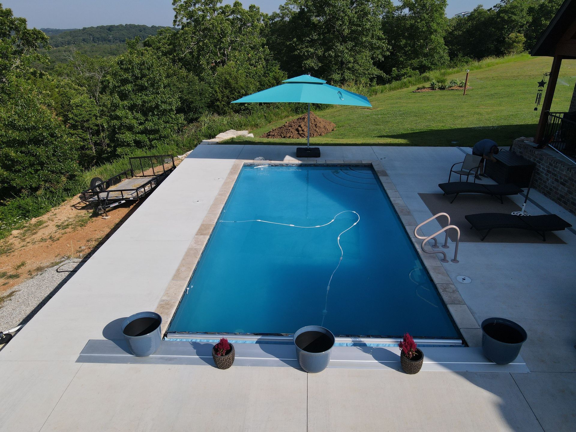 Swimming pool surrounded by concrete, umbrella, and view of trees.