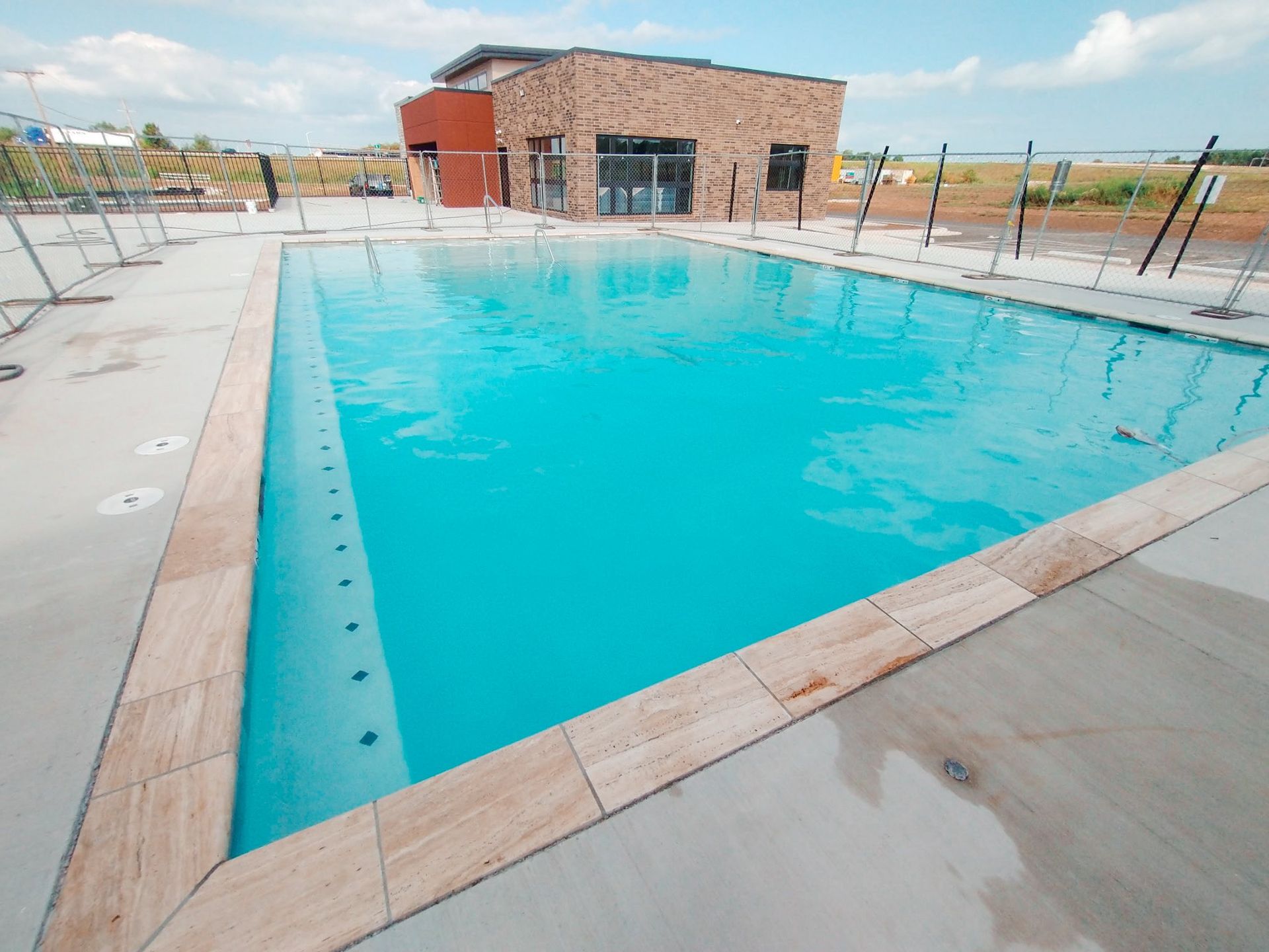Swimming pool with turquoise water, beige stone border, modern building in background. Sunny day.