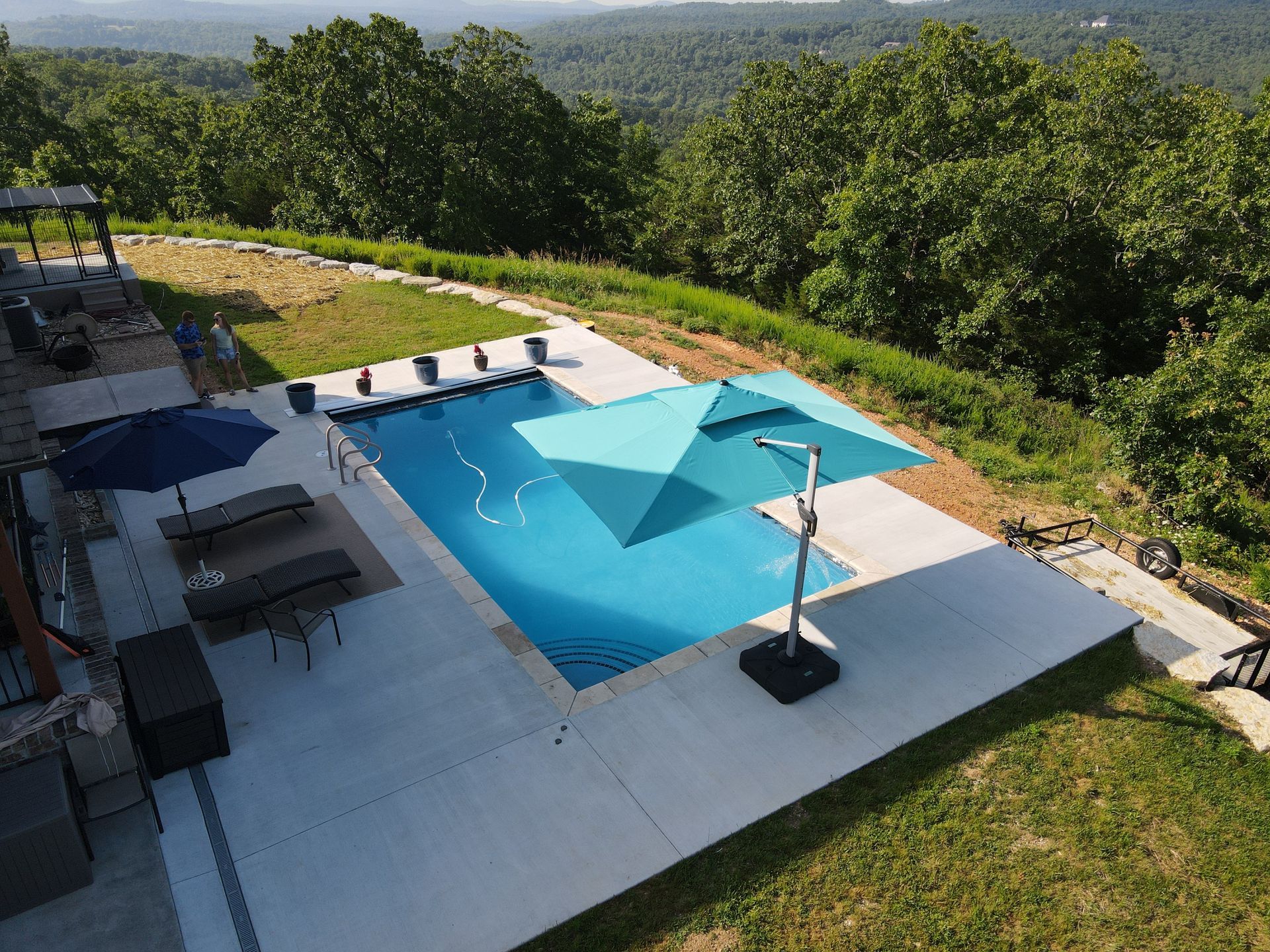 Swimming pool with teal umbrella on a sunny day overlooking green trees and rolling hills.