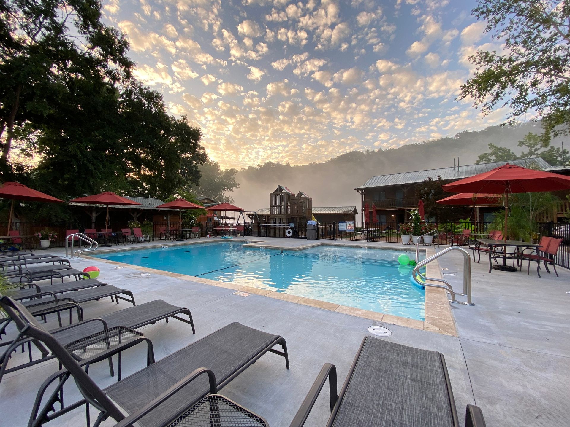 A pool with lounge chairs, red umbrellas, and a building under a cloudy sunrise.