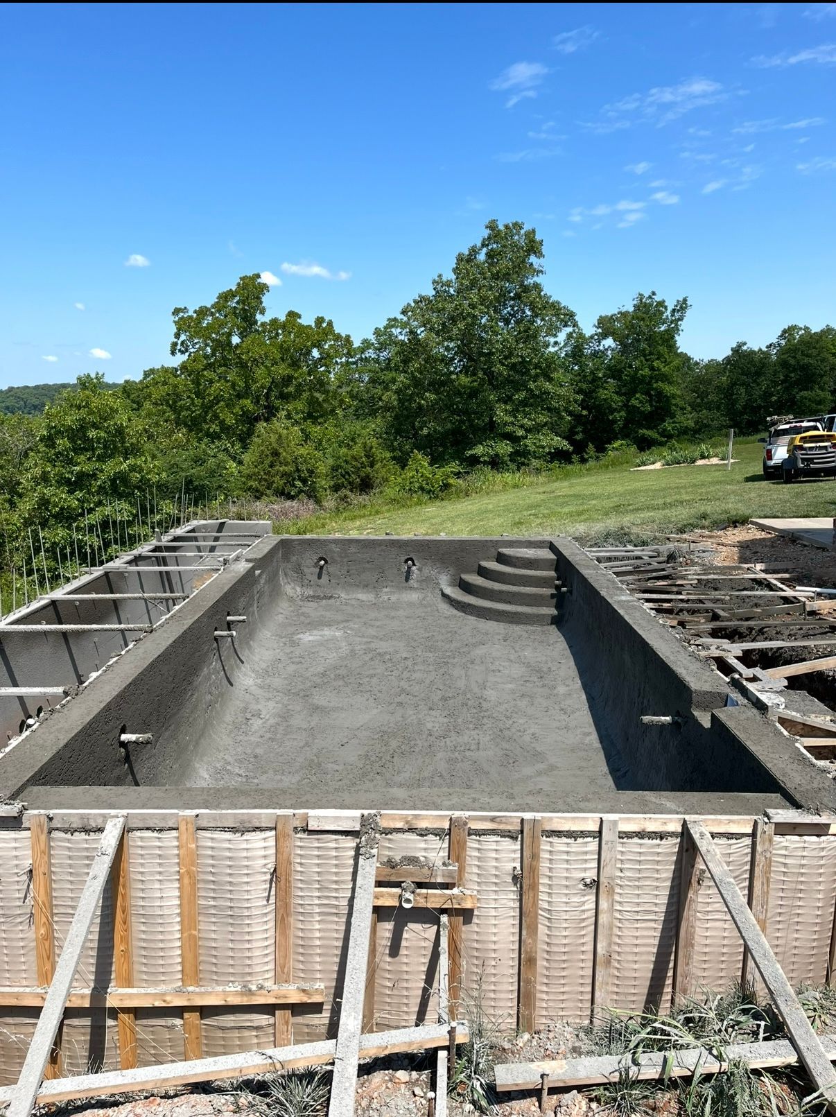 Concrete pool under construction, with steps, surrounded by wooden forms, and overlooking a green landscape under a blue sky.
