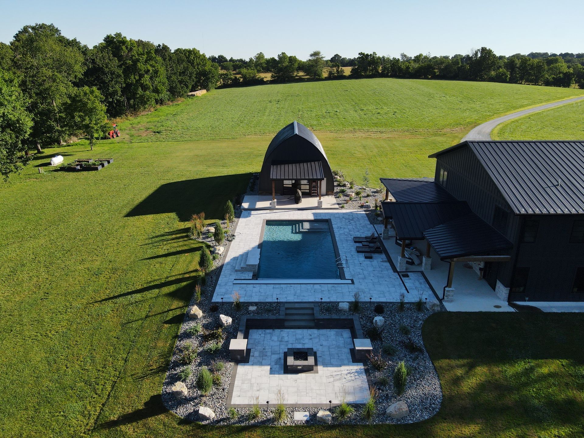 Pool and patio area next to a dark barn-style building on a grassy field.