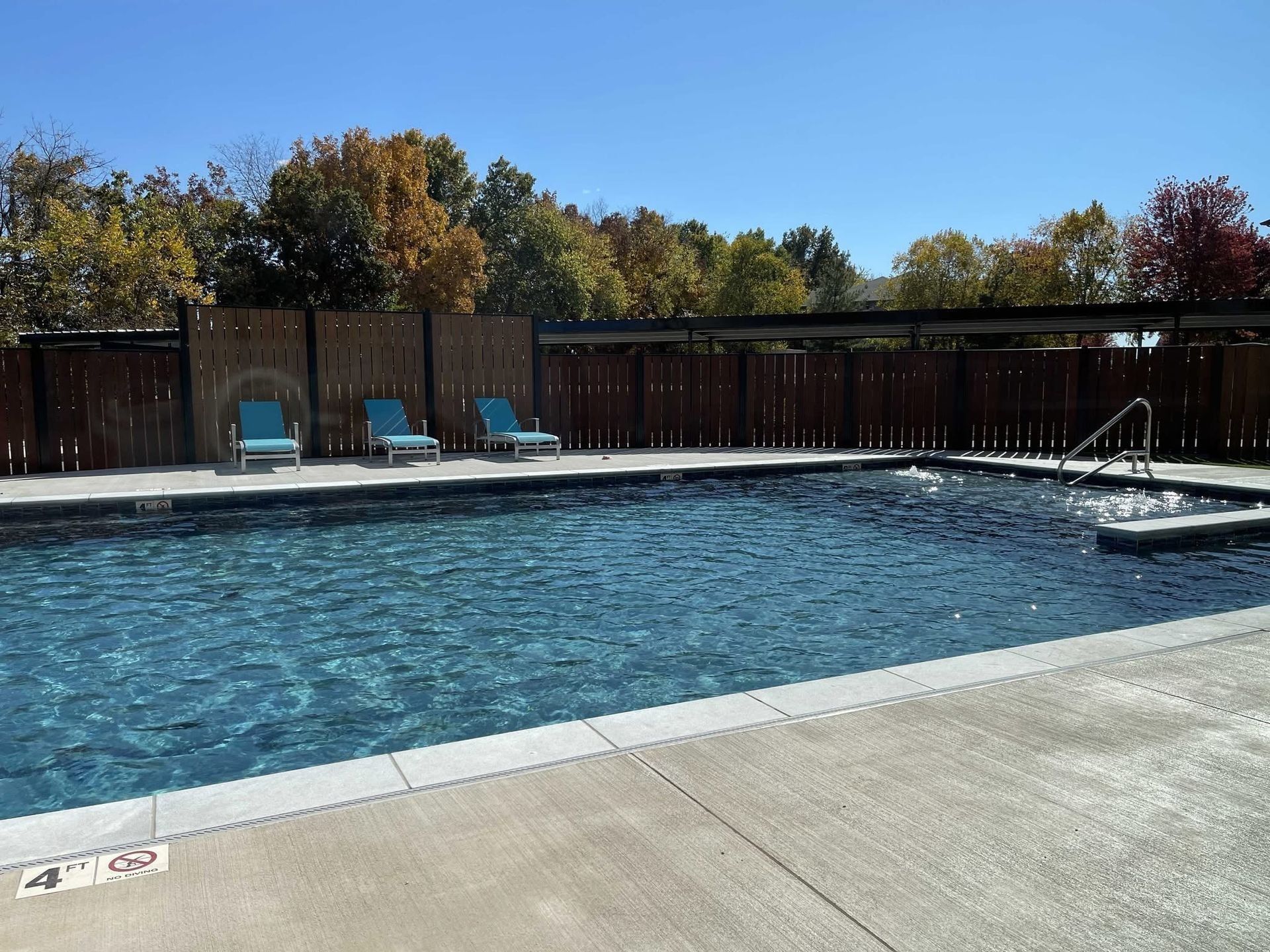 Outdoor swimming pool with three blue lounge chairs, surrounded by a wooden fence and trees with fall foliage.