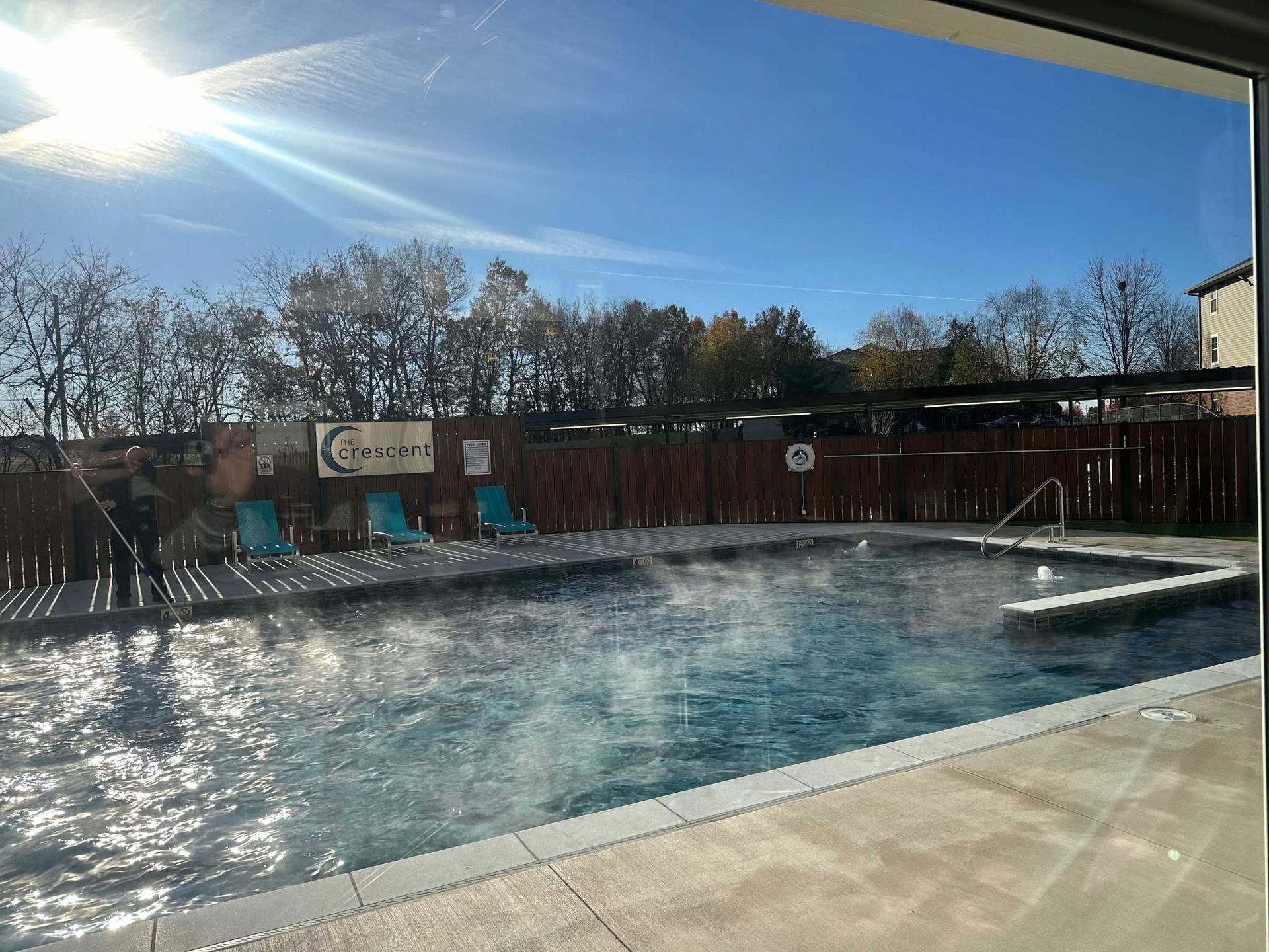 Pool with steam rising under a sunny blue sky. Lounge chairs along the side, fence in background.