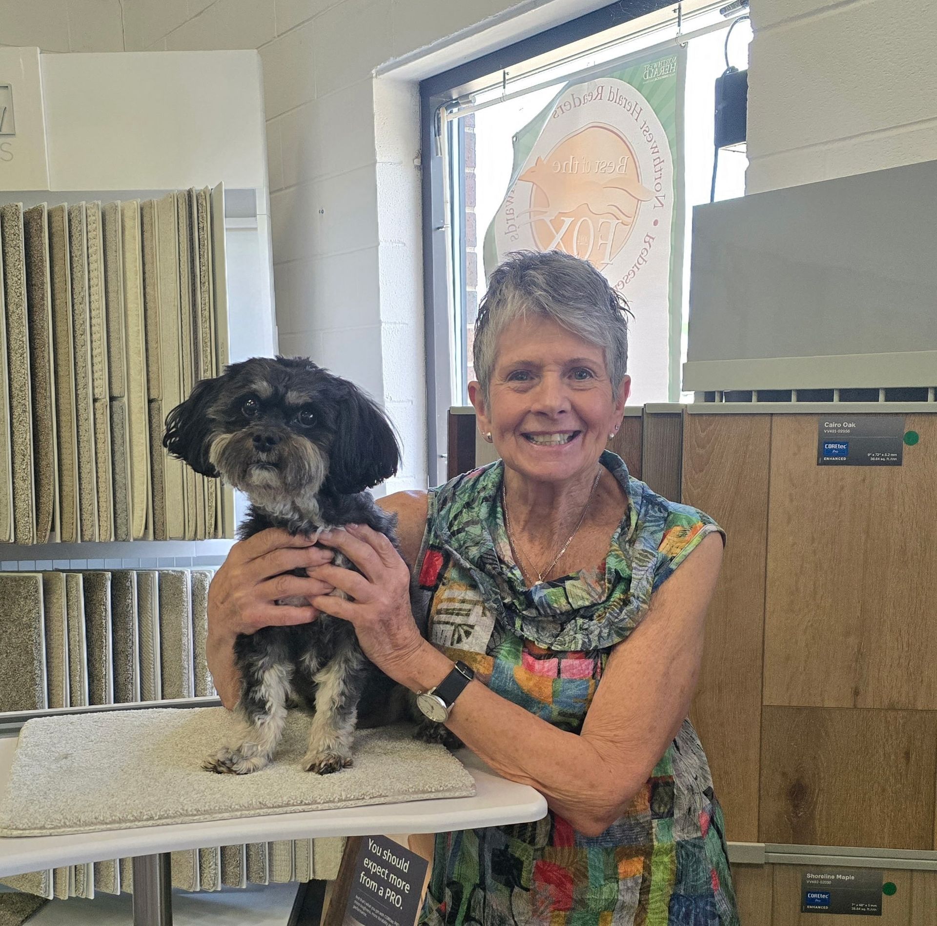 Woman smiles, holding small dog on a table in a flooring store.