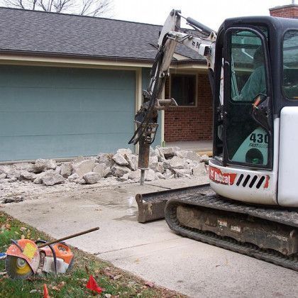 Mini excavator demolishing a concrete driveway beside a garage and house with debris piled nearby