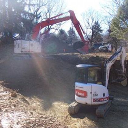 Two excavators on a dusty construction site, one red and one white, with trees in the background