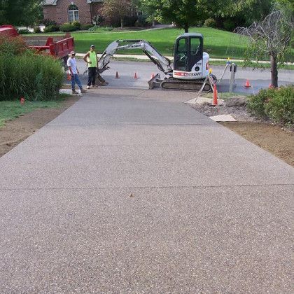 Workers repair a driveway with a small excavator and orange cones in a residential street