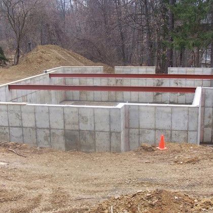 Concrete foundation walls under construction in a wooded area, with an orange traffic cone nearby.