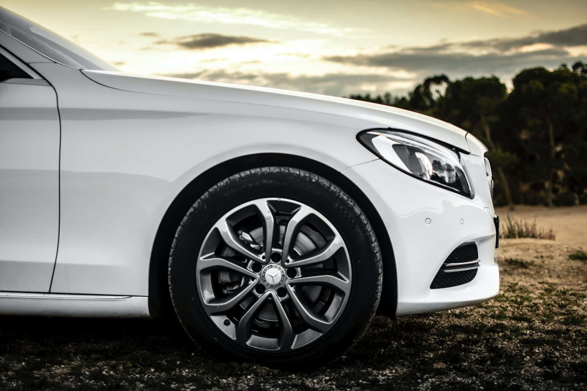 White Mercedes-Benz sedan, front wheel and headlight visible, parked outdoors on a cloudy day.