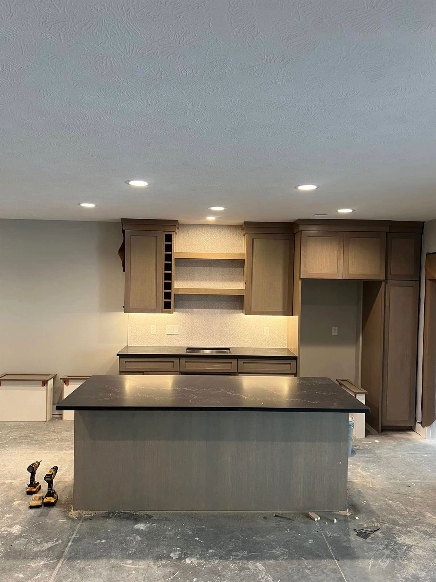 Kitchen with dark island, grey cabinets, and backsplash under construction.