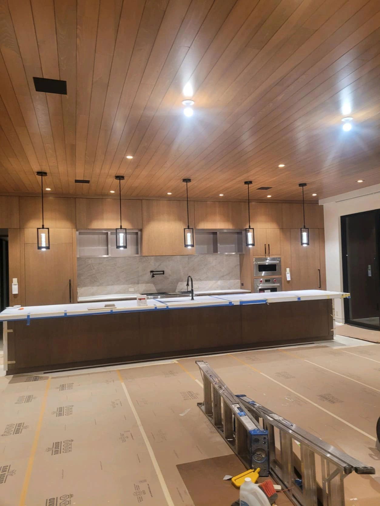 Modern kitchen under construction with wood cabinets and ceiling, island, pendant lights.