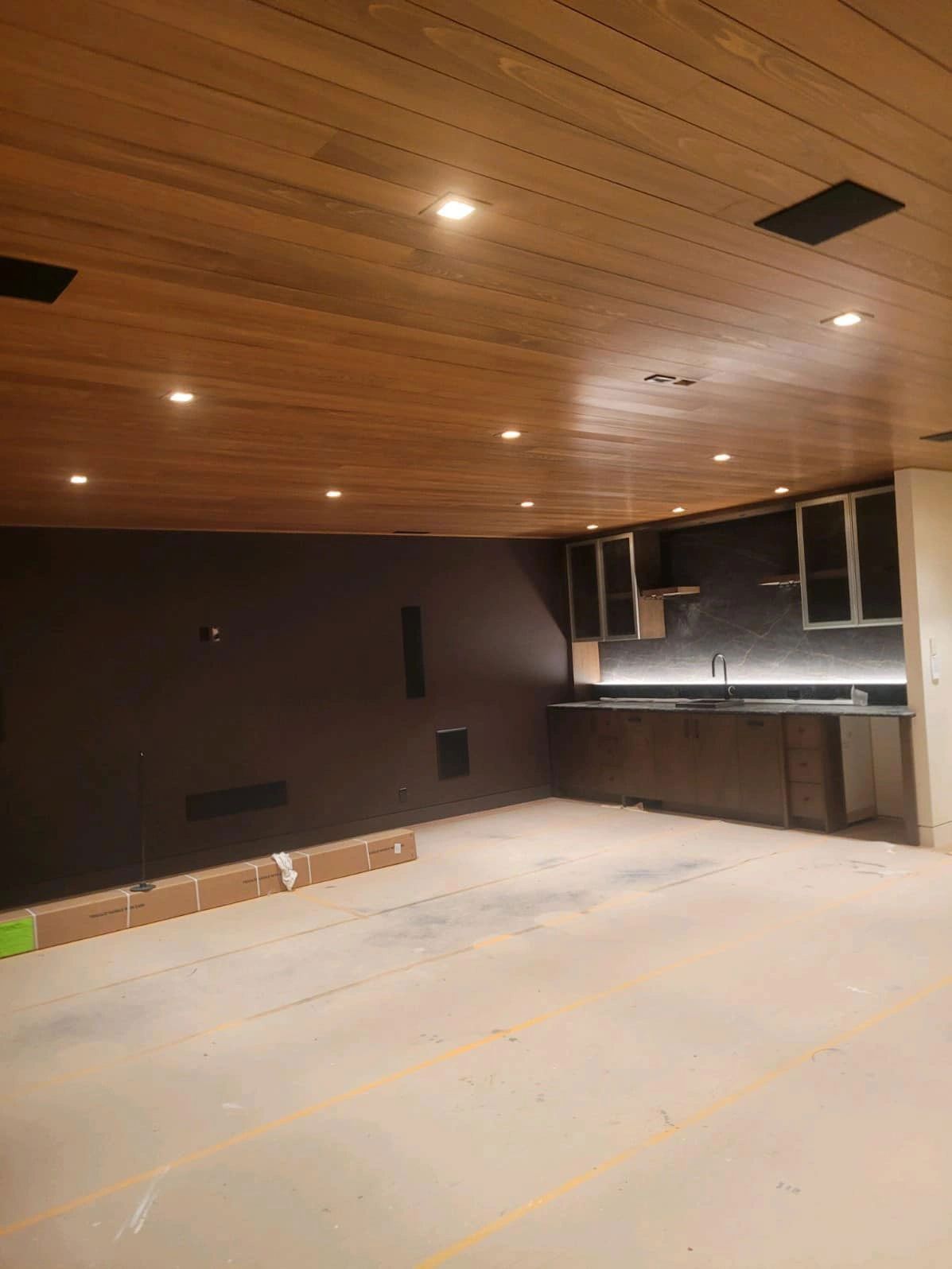 Empty room with wood ceiling, dark wall, small kitchen area, recessed lighting, and concrete floor.